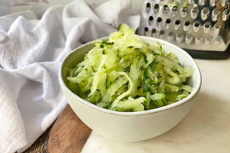 Horizontal image of shredded cucumber in a white bowl in front of a metal kitchen tool.