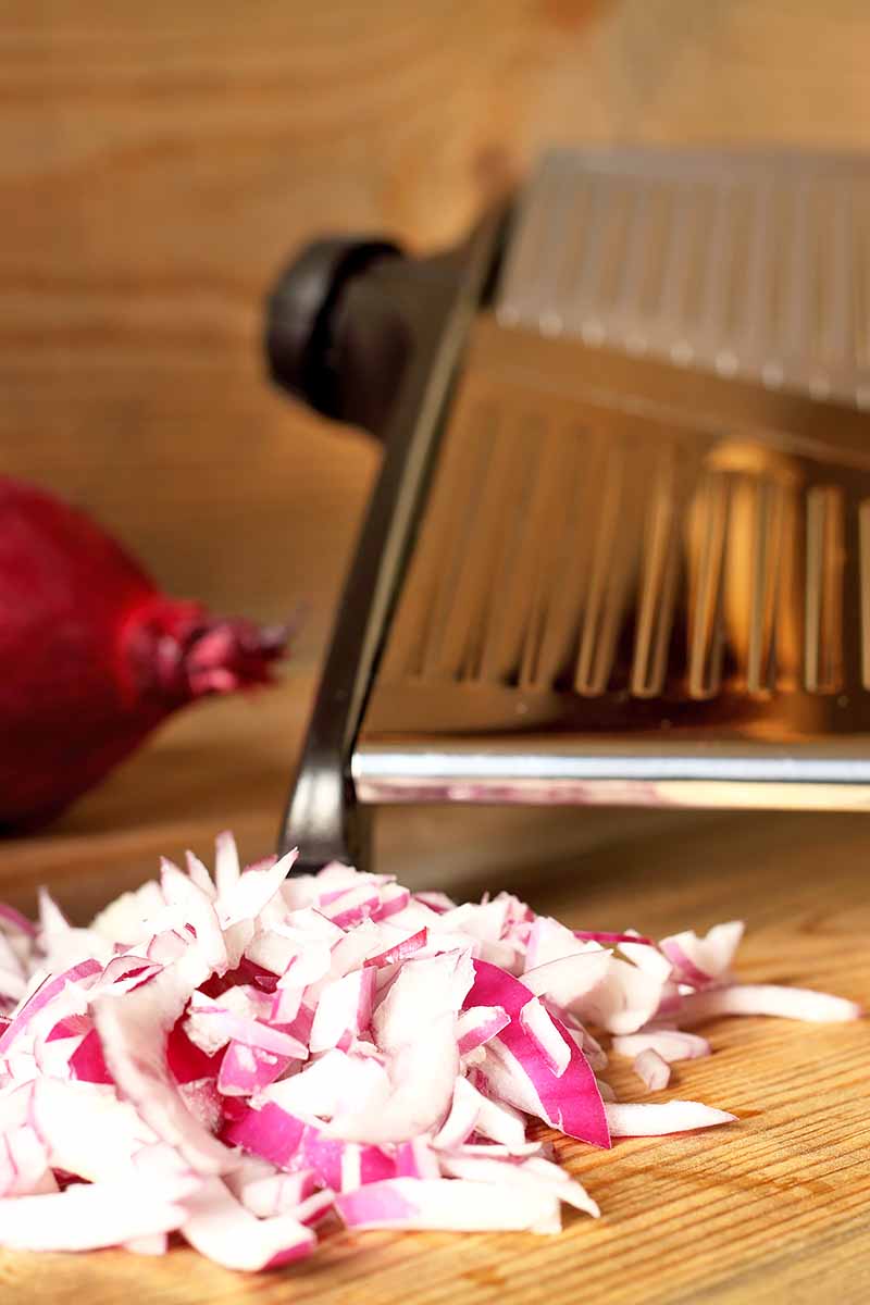 Vertical image of a metal kitchen gadget next to chopped red onions.