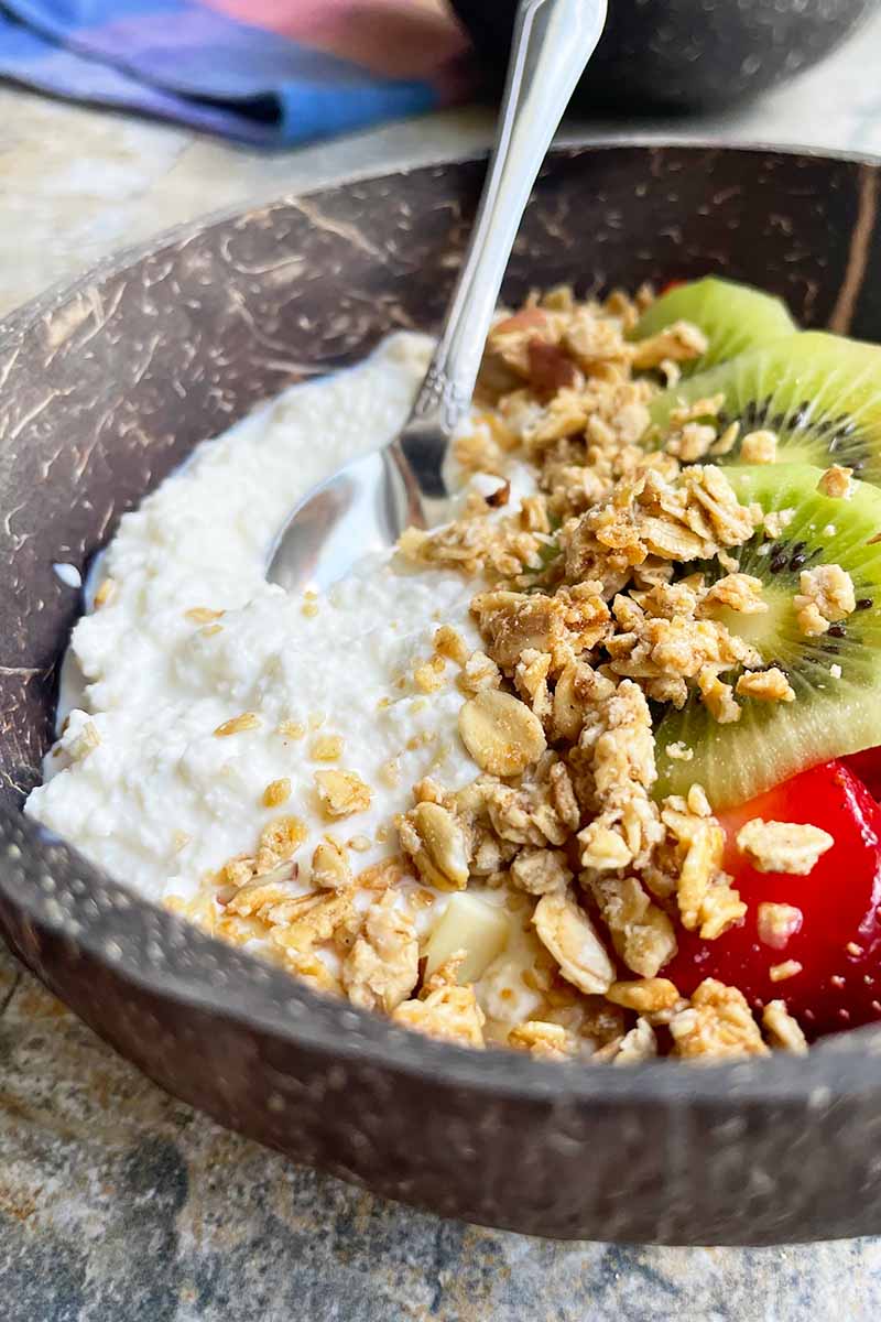 Vertical image of a wooden bowl filled with cottage cheese and garnishes of granola and sliced fresh fruit.
