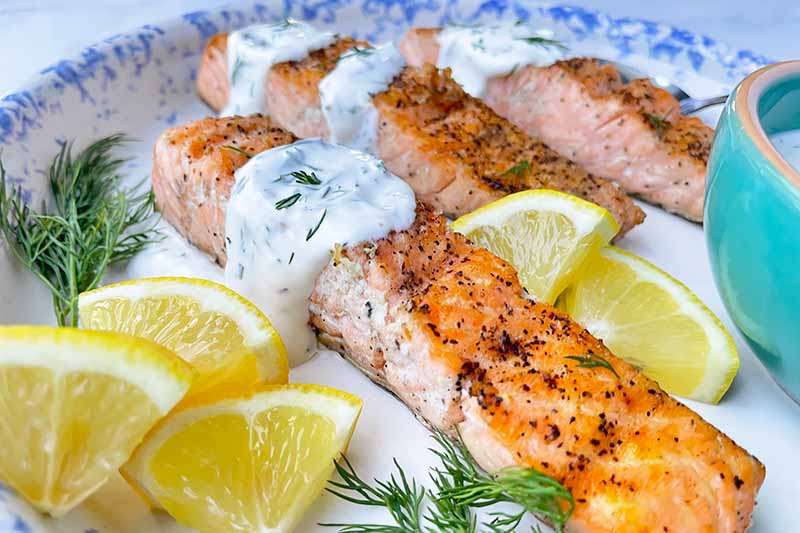 Horizontal close-up image of three salmon fillets on a plate topped with a creamy herb spread next to herbs, silverware, and lemon wedges.