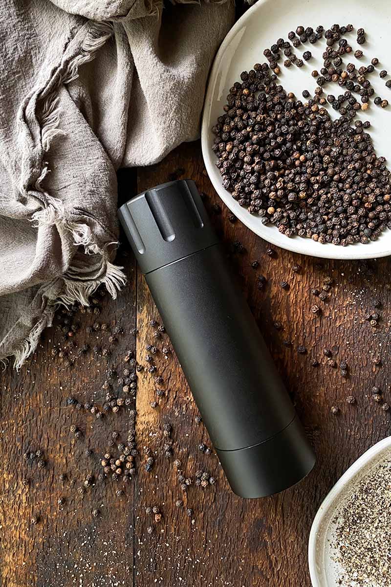 Vertical image of a black pepper grinder in the middle on a wooden table next to whole and ground spices and a tan towel.