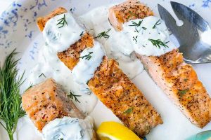 Horizontal image of three salmon fillets on a plate topped with a creamy herb spread next to herbs, silverware, and lemon wedges.