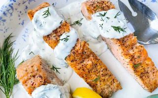 Horizontal image of three salmon fillets on a plate topped with a creamy herb spread next to herbs, silverware, and lemon wedges.