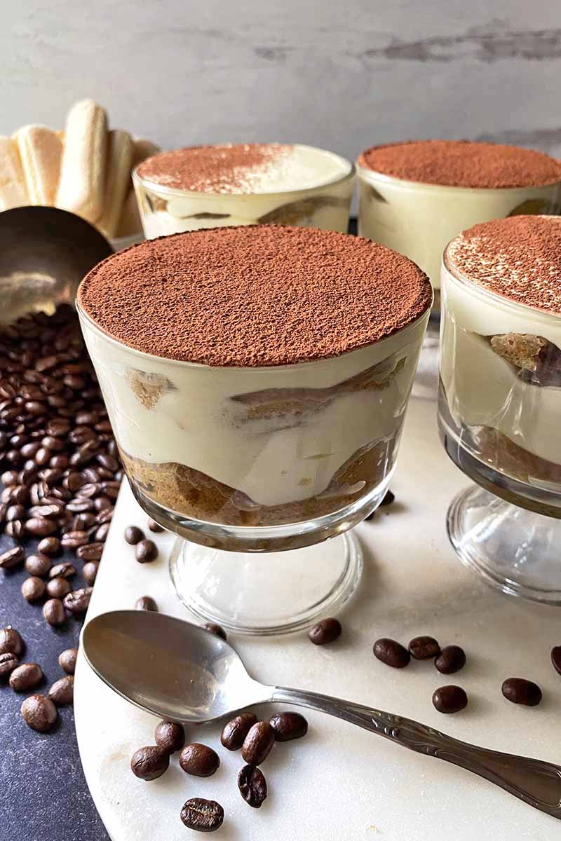 Vertical image of glass dishes filled with a trifle topped with cocoa powder on a marble slab next to a spoon and coffee beans.