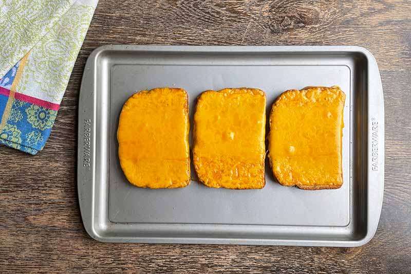 Horizontal image of melted orange cheese on slices of bread on a baking sheet.