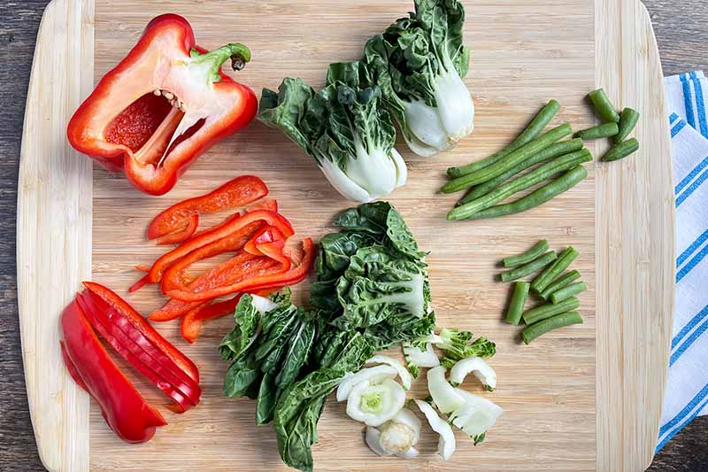 Horizontal image of bell peppers, green beans, and bok choy prepped on a wooden cutting board.