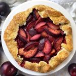 Horizontal top-down image of a large crostata with red fruit wedges next to a bowl of vanilla ice cream and metal forks on a white towel.