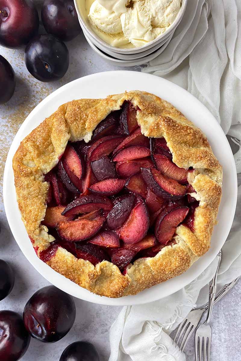 Vertical top-down image of a large crostata with red fruit wedges next to a bowl of vanilla ice cream and metal forks on a white towel.