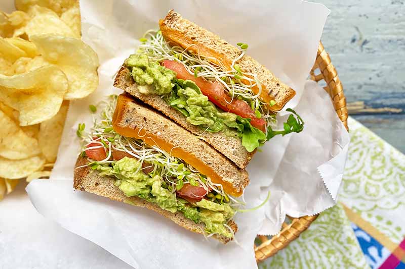 Horizontal image of a halved veggie sandwich in a paper-lined basket next to potato chips.