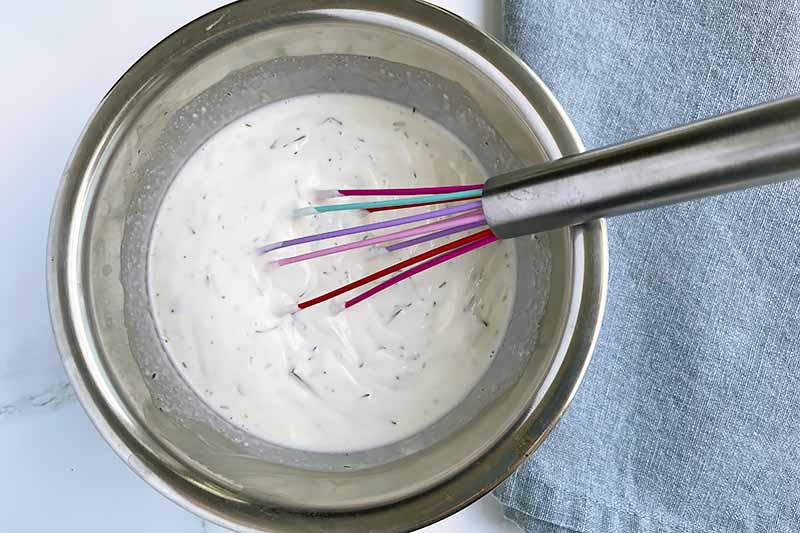 Horizontal image of a whisk in a yogurt and herb mixture in a metal bowl.