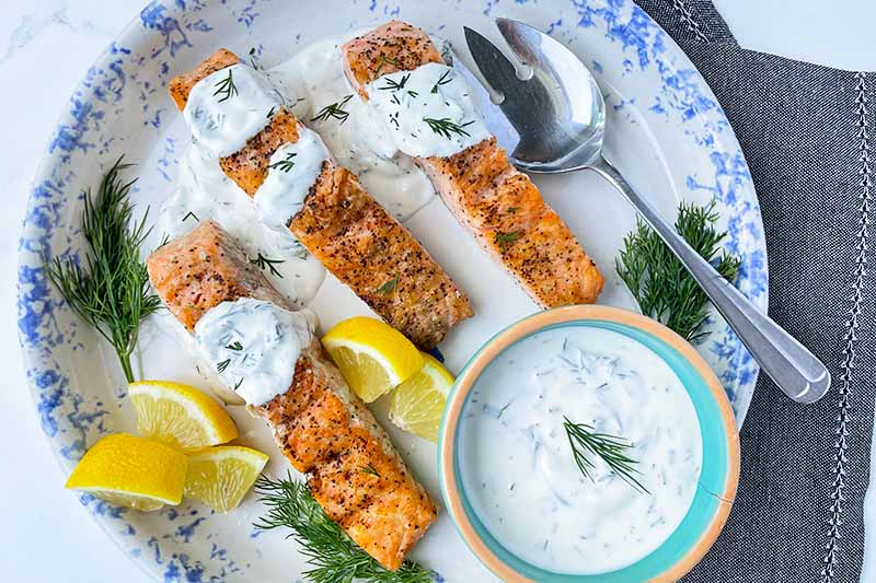 Horizontal top-down image of three salmon fillets on a plate topped with a creamy herb spread next to silverware and lemon wedges.