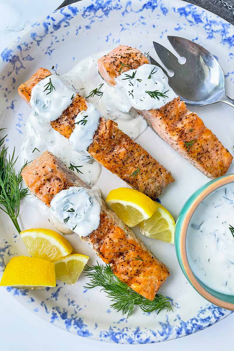 Vertical top-down image of three salmon fillets on a plate topped with a creamy herb spread next to silverware and lemon wedges.