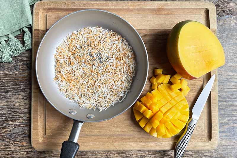 Horizontal image of toasted coconut shreds and cut mango on a cutting board.