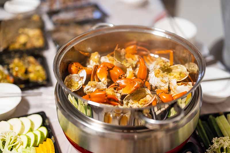 Horizontal image of steaming assorted shellfish in a large pan surrounded by assorted vegetables on the table.