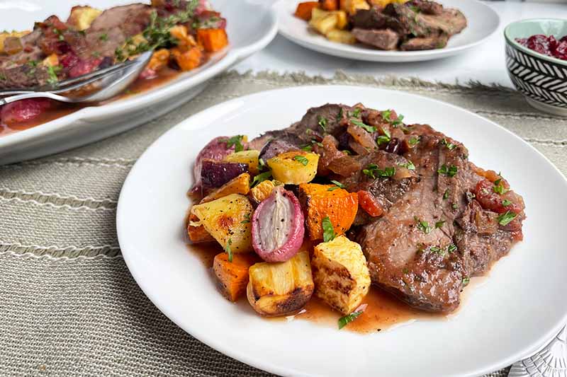 Horizontal image of white plates and a platter with servings of sliced beef in a sauce next to roasted assorted vegetables.