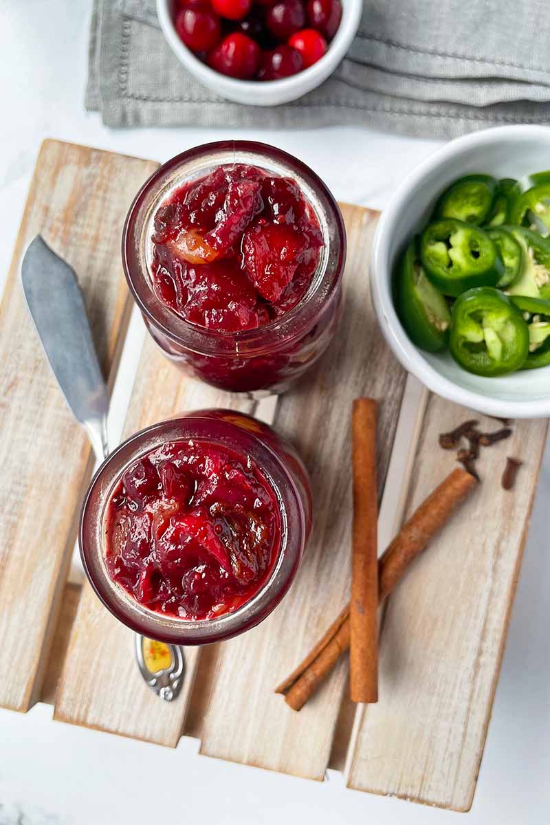 Vertical top-down image of two jars full of a thick fruit spread on a wooden board next to cinnamon and a bowl of sliced jalapenos.