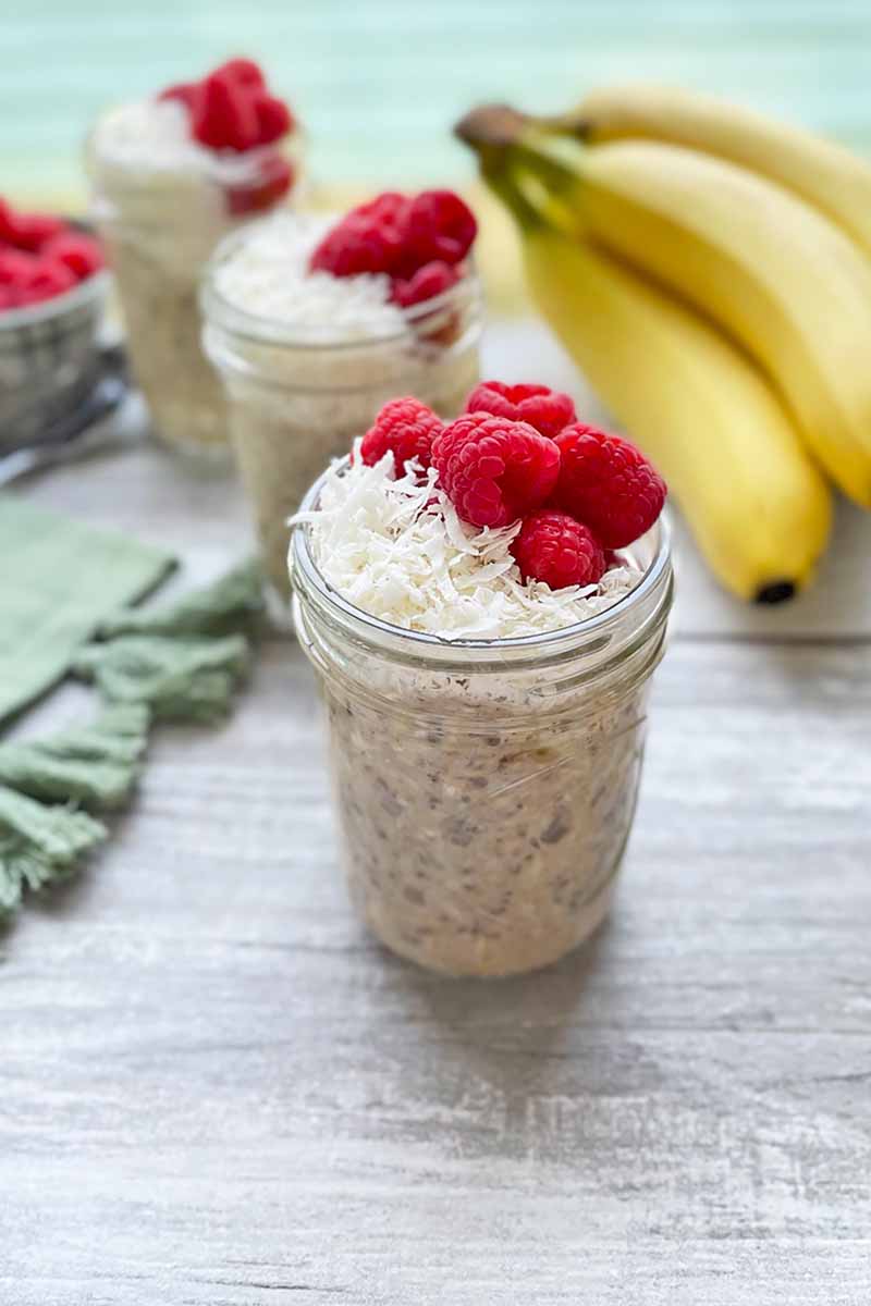 Vertical image of three glass mason jars filled with shredded coconut and fresh raspberries next to bananas.