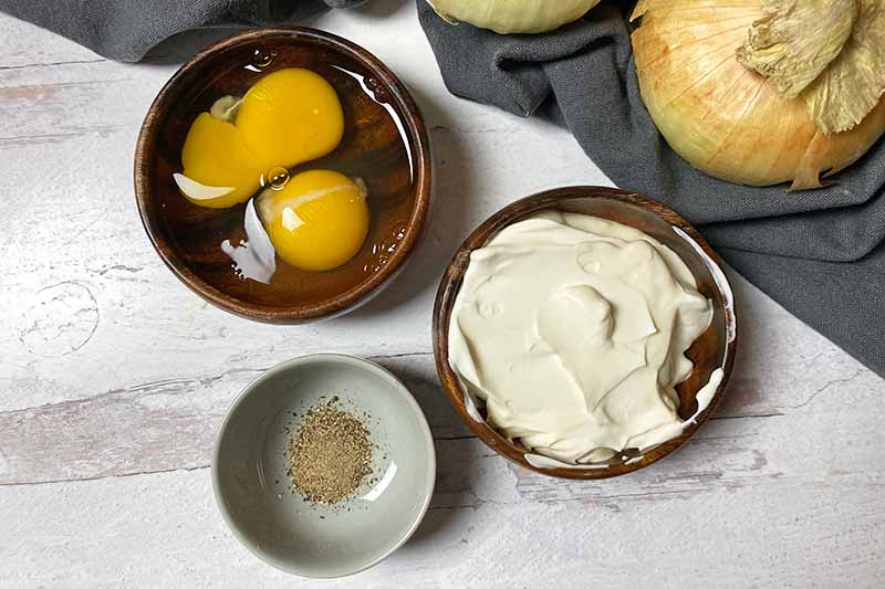 Horizontal image of a bowl of raw eggs, a bowl of creme fraiche, and a bowl of black pepper.