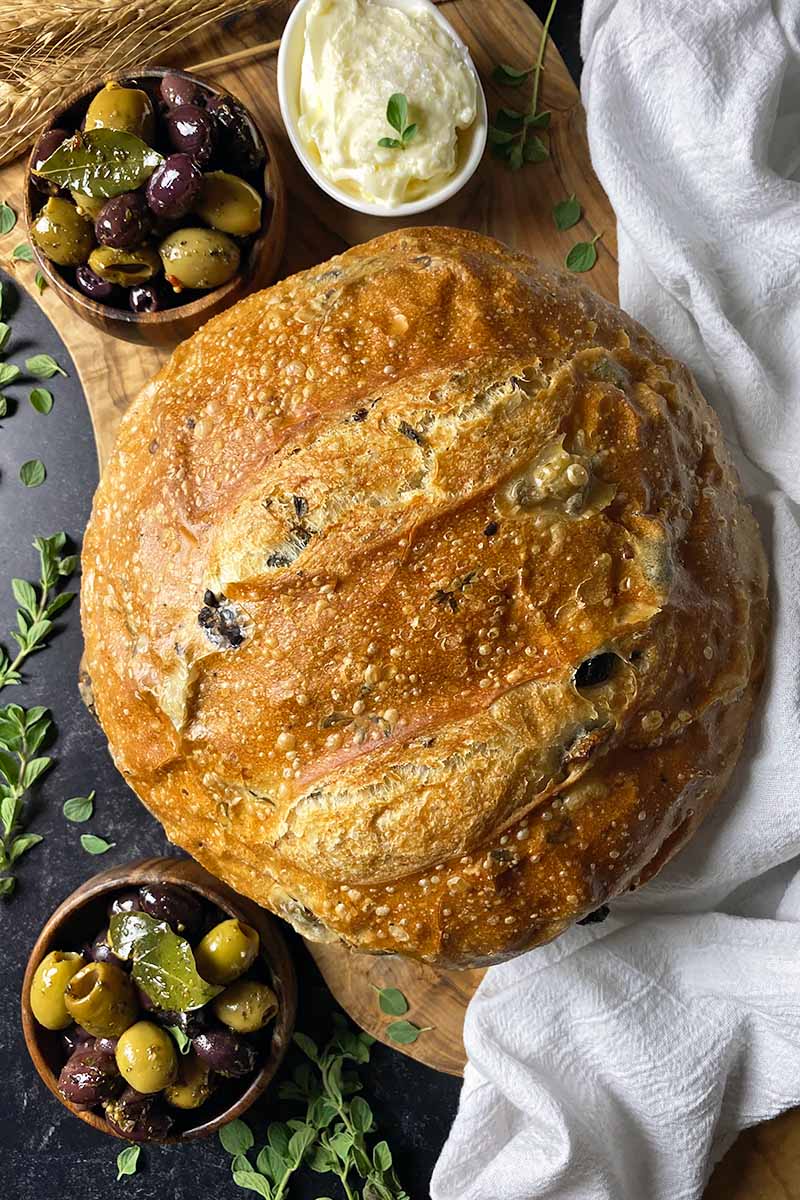 Vertical top-down image of a whole loaf of bread next to olives, whipped butter, and a white towel.