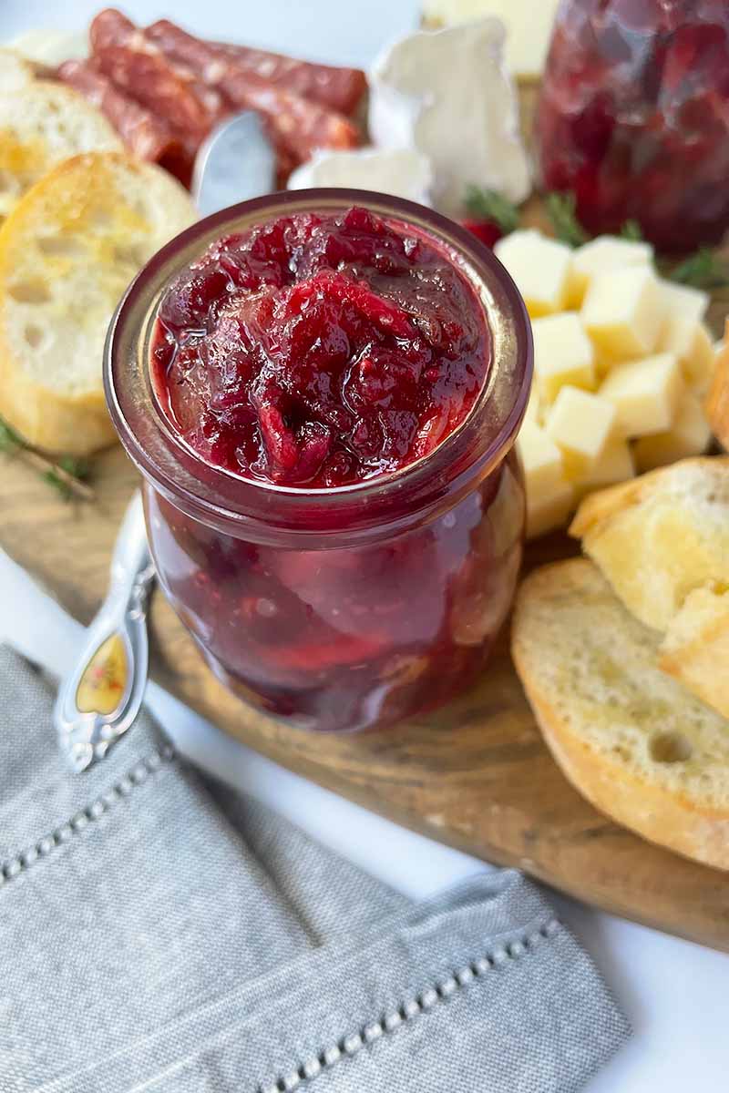 Vertical image of a glass jar filled with a chunky red fruit spread on a cheeseboard.