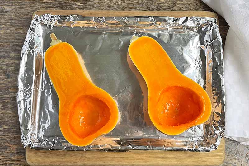 Horizontal image of oiled halves of a butternut on a lined baking sheet.