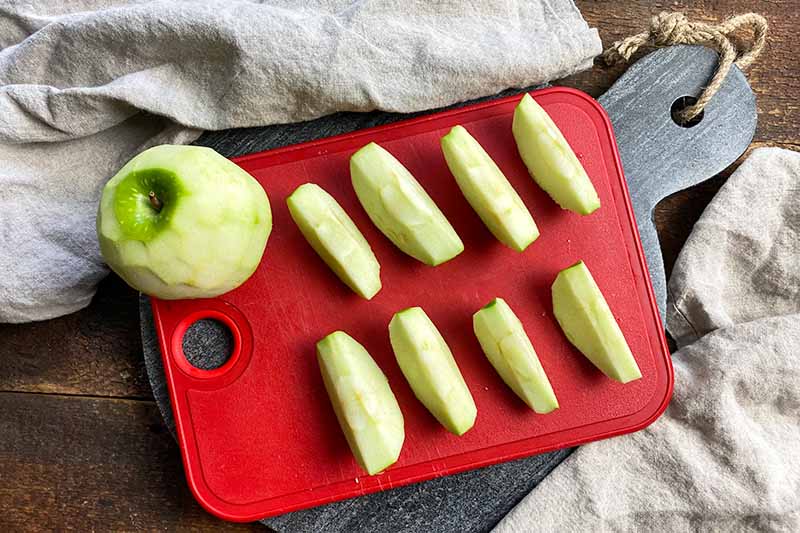 Horizontal image of wedges and a whole peeled apple on a red cutting board.