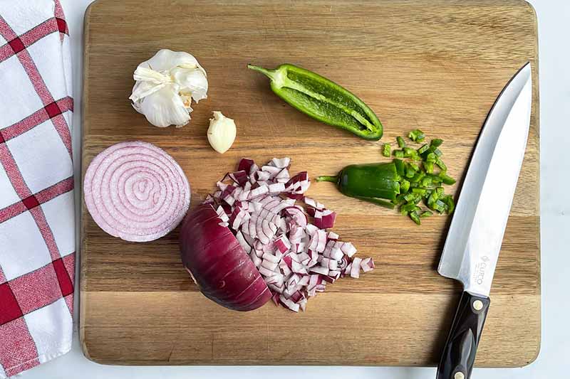 Horizontal image of chopped onions, jalapenos, and garlic on a wooden cutting board next to a knife.