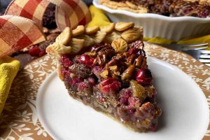 Horizontal image of a slice of a fruit and nut pastry on a white plate next to plaid fabric and a casserole dish.