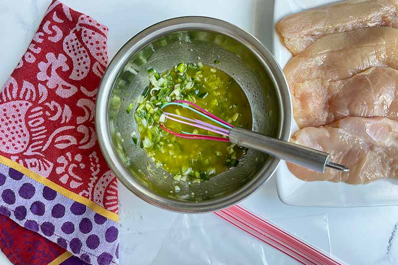 Horizontal image of whisking together a green sauce in a metal bowl next to raw meat pieces on a white platter.