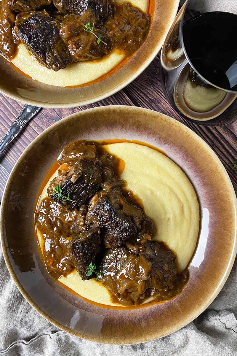 Vertical top-down image of two brown bowls filled with creamy polenta and stewed meat next to a glass and tan napkin.