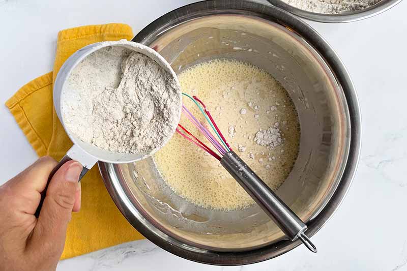 Horizontal image of adding dry ingredients into a batter in a metal bowl with a whisk.