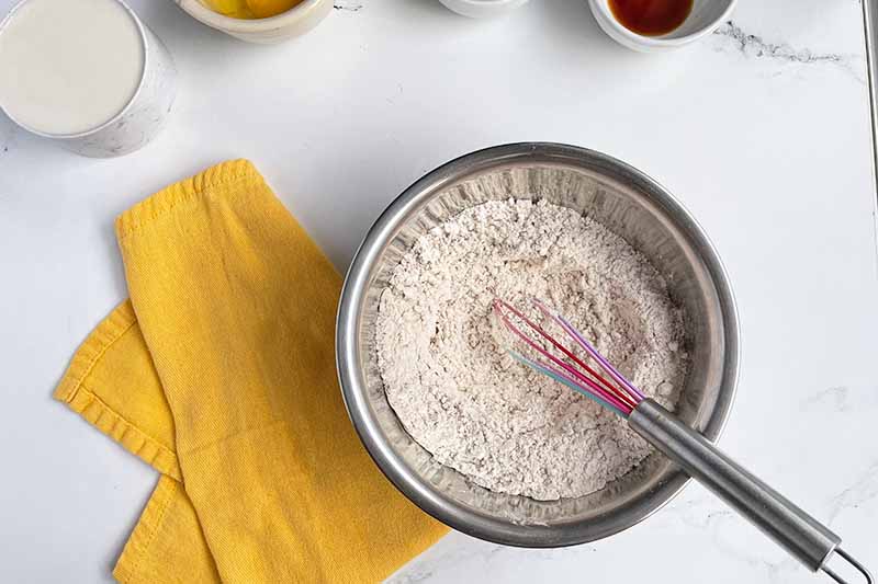 Horizontal image of whisking together dry ingredients in a metal bowl on a yellow towel.