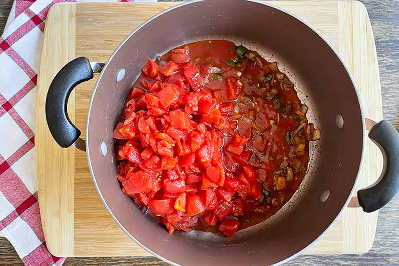 Horizontal image of a pot with aromatics and chopped tomatoes cooking.