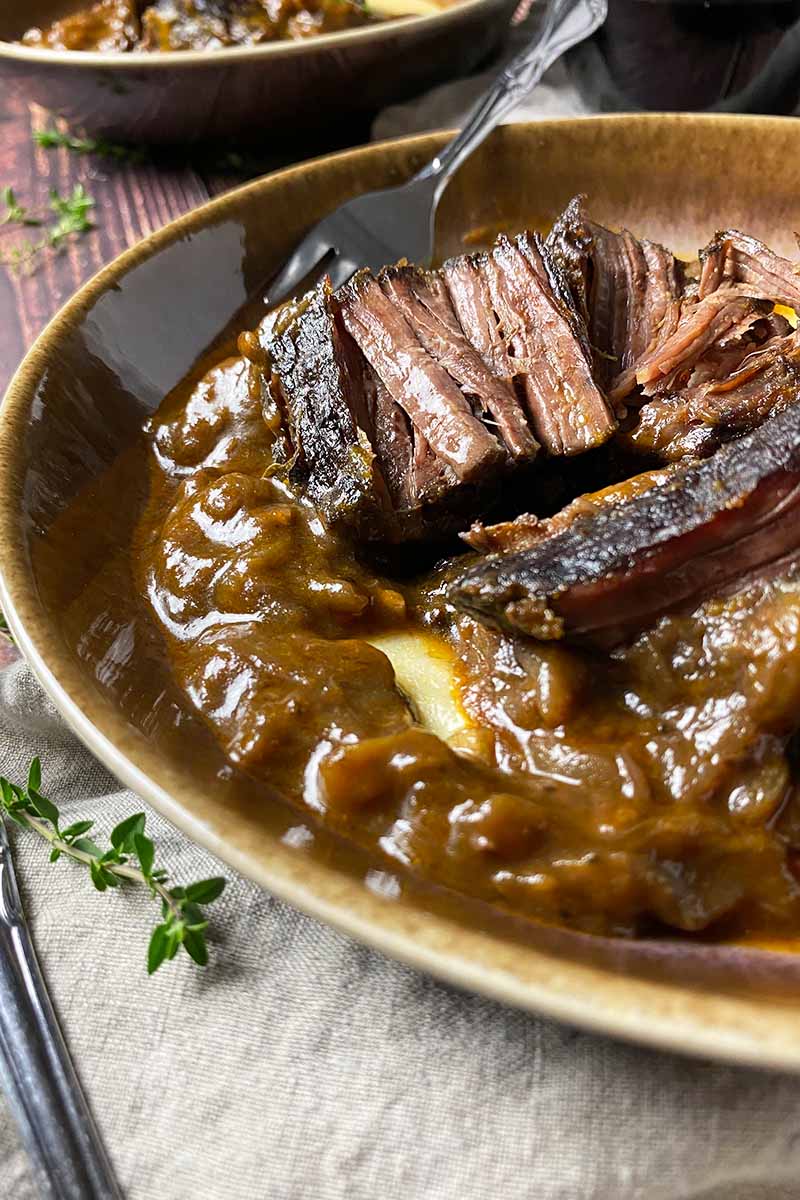 Vertical image of large shreds of stewed meat in a brown bowl over polenta next to thyme sprigs and a tan towel.