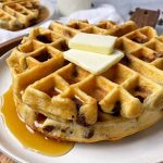 Horizontal image of a stack of waffles topped with butter slices with syrup on a white plate on a wooden platter in front of glasses of milk.