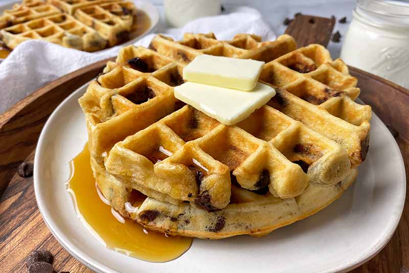Horizontal image of a stack of waffles topped with butter slices with syrup on a white plate on a wooden platter in front of glasses of milk.