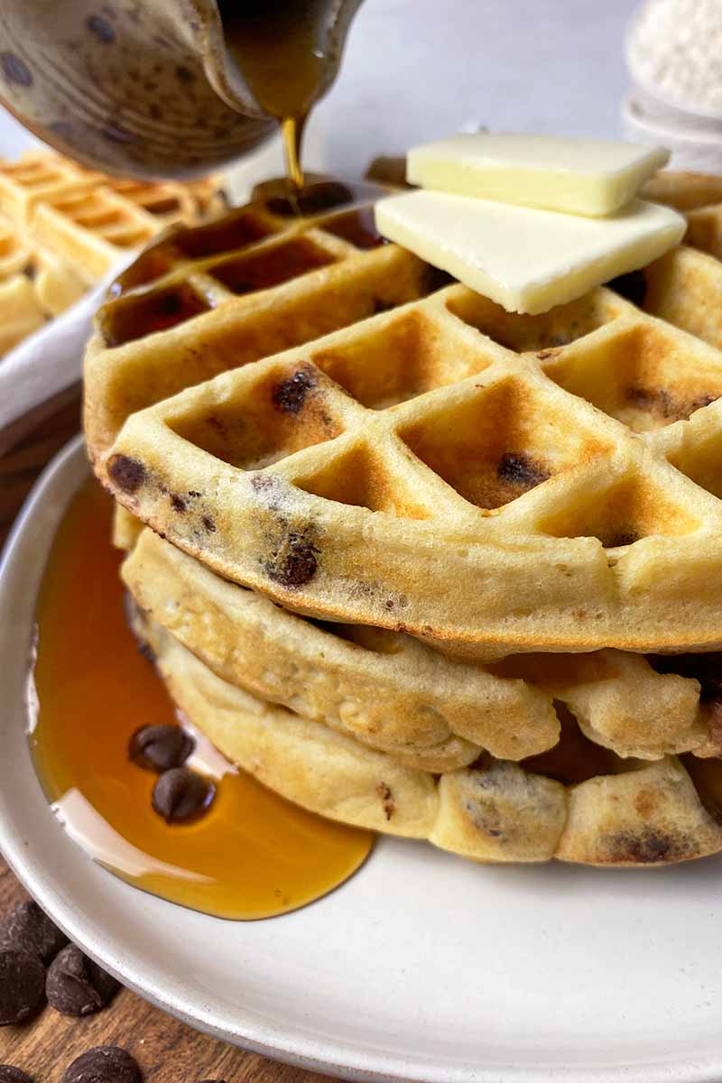 Vertical image of pouring maple syrup over a stack of Belgian waffles topped with slices of butter on a white plate.