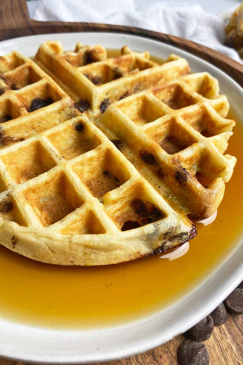 Horizontal image of a waffle over maple syrup on a white plate.