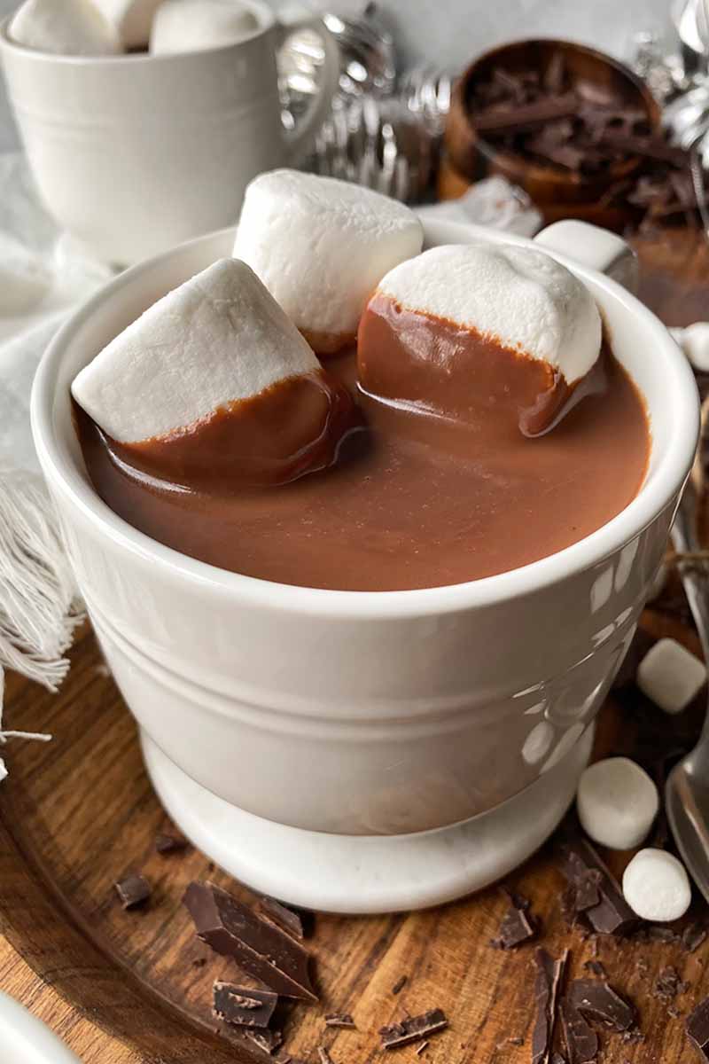 Vertical image of two mugs filled with a dark brown creamy beverage topped with jumbo marshmallows on a wooden board.