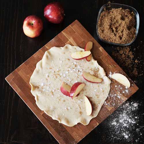 Image of a wooden board underneath a pastry surrounded by apples and cinnamon sugar.