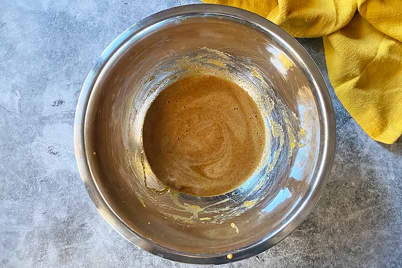 Horizontal image of a light brown liquid mixture in a metal bowl next to a yellow towel.