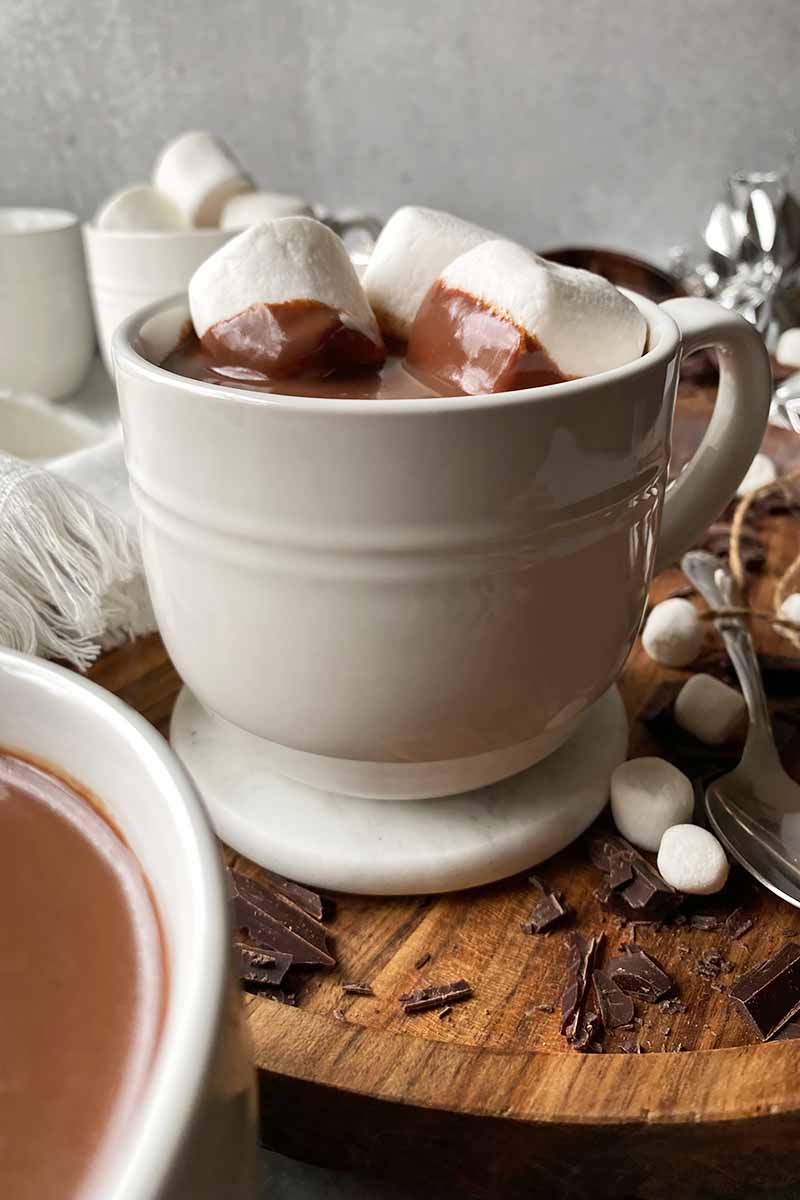 Vertical image of a large white mug filled with a creamy dark brown drink topped with marshmallows on a wooden board.