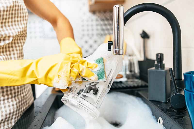 Horizontal image of a person cleaning a plastic container in a sink with a sponge while wearing yellow gloves.