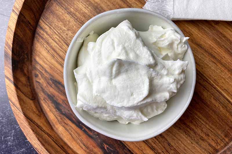 Horizontal image of whipped cream in a white bowl on a wooden platter.