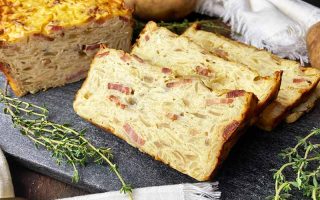 Horizontal image of slices of a savory cake with pieces of thick-cut bacon on a dark slate board next to thyme leaves.