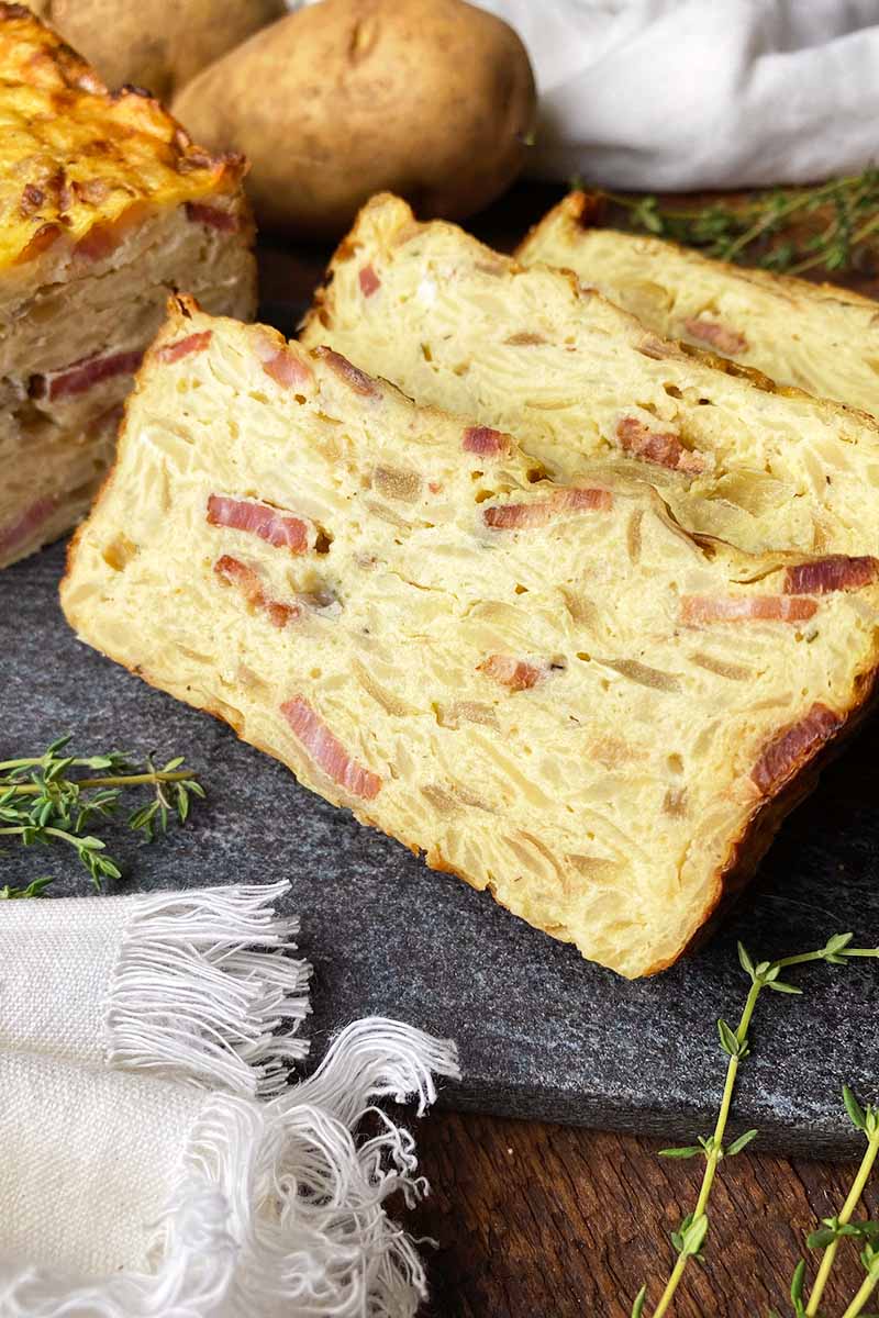 Vertical image of slices of a savory cake with pieces of thick-cut bacon on a dark slate board next to thyme leaves.