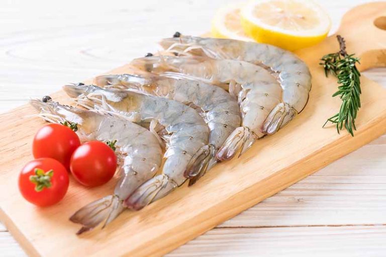 Horizontal image of raw crustaceans on a wooden board next to tomatoes, lemons, and herbs.