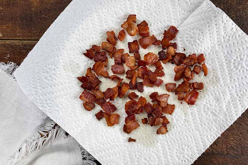 Horizontal image of draining chopped cooked crispy bacon pieces on a plate lined with a paper towel.