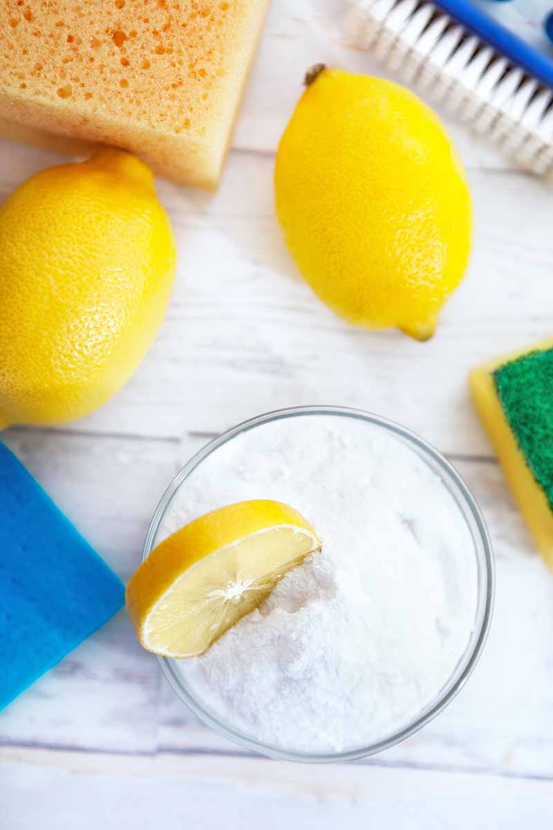 Vertical image of lemons and a bowl of baking soda next to sponges on a white surface.