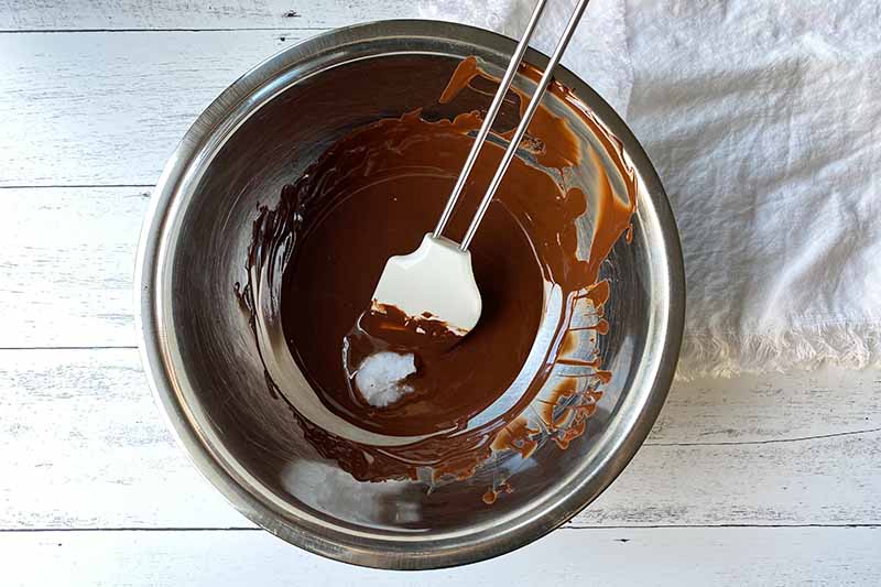 Horizontal image of melted chocolate and a tablespoon of coconut oil in a bowl stirred with a spatula.
